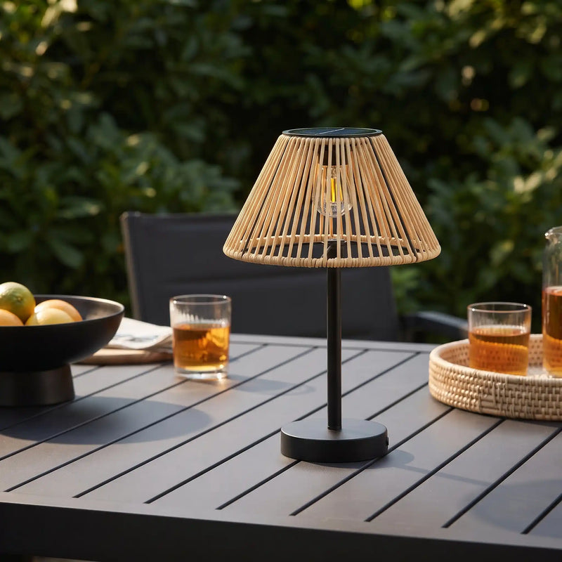 A modern table lamp with a black metal stand and a woven natural-fiber conical shade, placed on a dark outdoor patio table with drinks and greenery in the background.