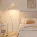 A modern floor lamp with a pleated white shade and curved arm stands beside a neatly made bed in a softly lit bedroom. The lamp is turned on, casting a warm glow, and the room has cream bedding, pillows, a small side table, and framed wall art.