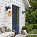 Two outdoor solar wall sconces with woven natural wood shades and black mounts, shown on either side of a blue front door on a white-shaded house exterior.