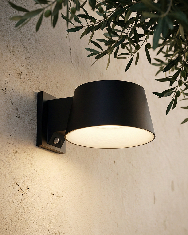 A modern matte black outdoor wall sconce mounted on a textured beige stucco wall, casting a warm glow upwards and downwards, with green olive branches hanging from above.