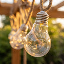 Close-up of clear Edison bulb with copper filament inside, suspended on jute rope; soft bokeh highlights waterproof design and vintage charm.