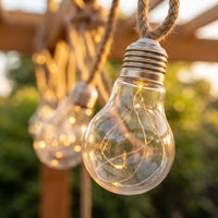 Close-up of clear Edison bulb with copper filament inside, suspended on jute rope; soft bokeh highlights waterproof design and vintage charm.