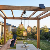 A wooden pergola with three hanging woven pendant lights in an outdoor backyard setting. A hand holds a black remote control in the foreground, and a small solar panel is mounted on the top beam.