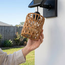 Close-up of hand installing PT0909 solar wall light: woman holding woven PE rattan shade beneath black mounting bracket with integrated solar panel — highlights tool-free assembly and natural material detail.