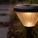 A close-up of a solar pathway light glowing outdoors at dusk, with a ribbed clear glass shade and a black top and post, set against a blurred garden background.