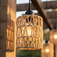 A cylindrical pendant light with a natural woven rattan or wicker shade, hanging outdoors under warm evening light near wooden beams.