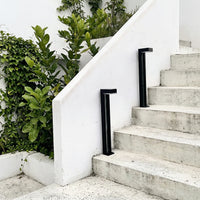 Two black anodised aluminium square solar bollards (Nivra PT0724) positioned alongside white concrete outdoor steps, with a white rendered wall and lush green foliage in the background, showcasing the lights in a real outdoor landscape setting.