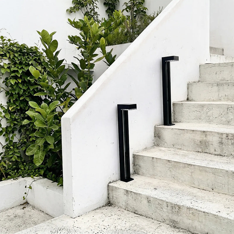 Two black anodised aluminium square solar bollards (Nivra PT0724) positioned alongside white concrete outdoor steps, with a white rendered wall and lush green foliage in the background, showcasing the lights in a real outdoor landscape setting.