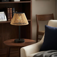 A table lamp with a black metal base and a natural bamboo slatted shade sits on a round wooden side table next to a beige armchair with a navy blue pillow in a cozy living room setting.