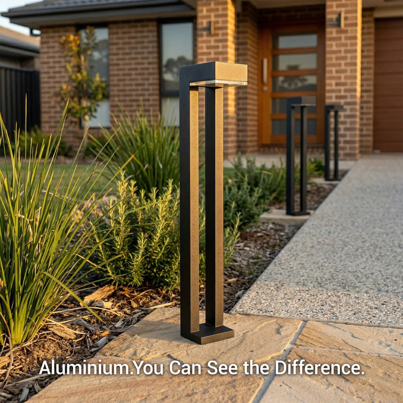 Outway PT0724 Nivra aluminium solar bollard light architectural detail daytime golden hour, black square column with brushed gold L-head, garden path