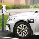 A white electric vehicle plugged into a charging station outdoors, with the charging cable connected to the car's charging port. The scene is set on a paved surface with grass and a blurred background.