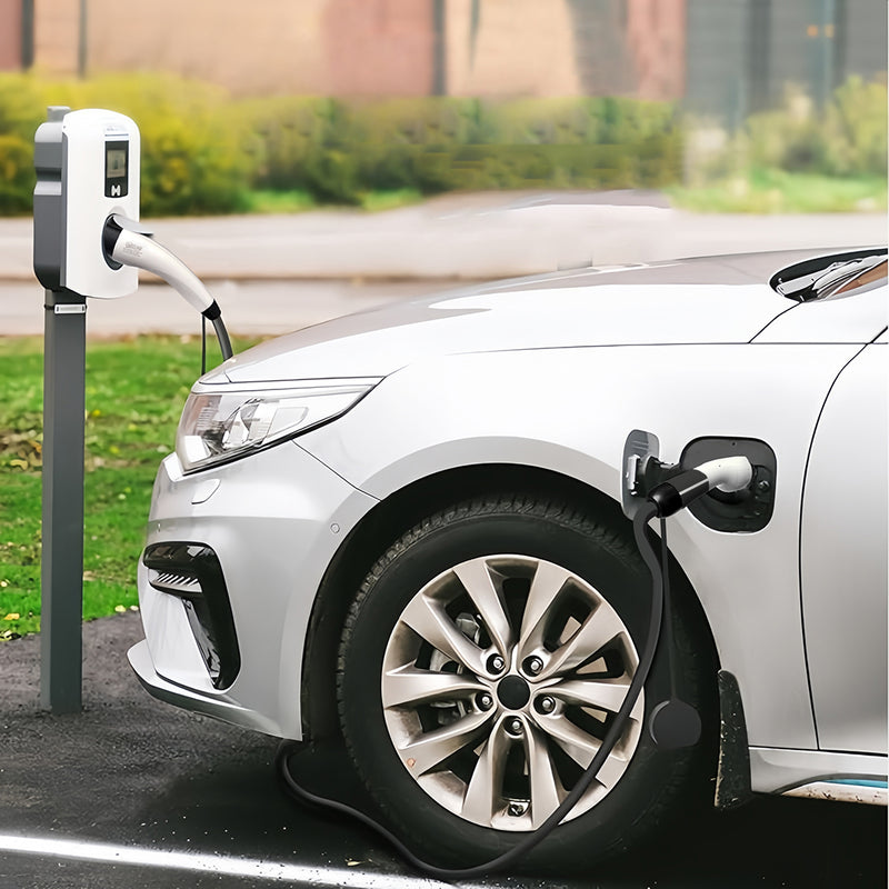 A white electric vehicle plugged into a charging station outdoors, with the charging cable connected to the car's charging port. The scene is set on a paved surface with grass and a blurred background.