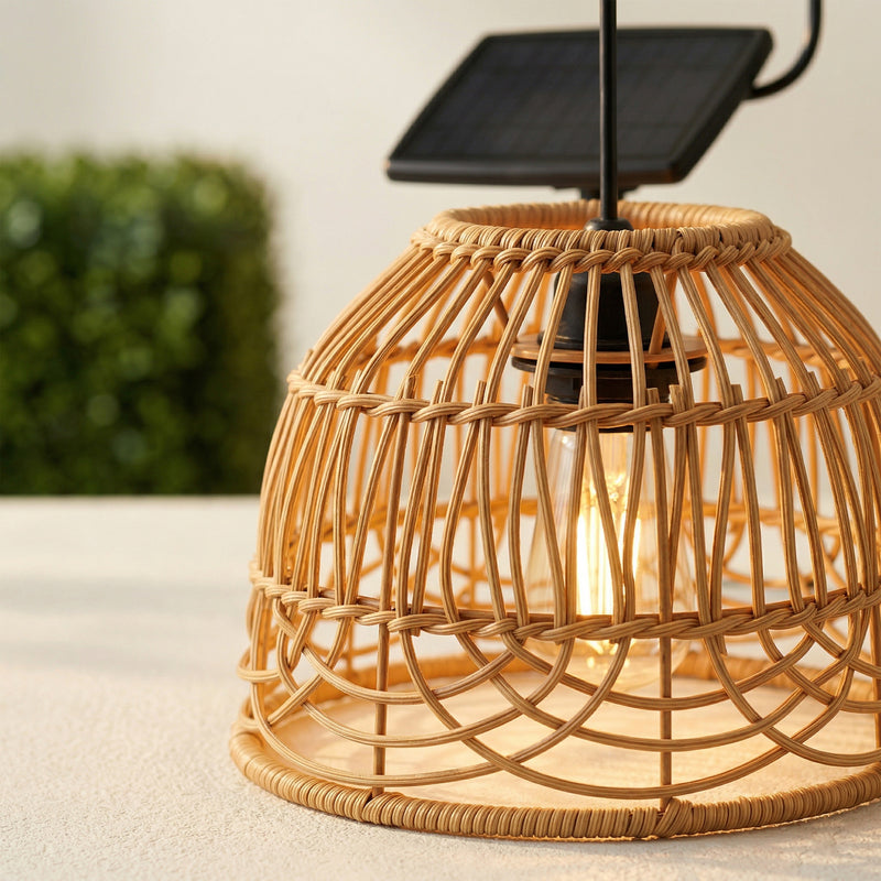 A close-up of a solar-powered pendant light with a natural woven rattan cage shade, glowing warm bulb, and a small black solar panel on a metal arm, set against a softly blurred outdoor background.
