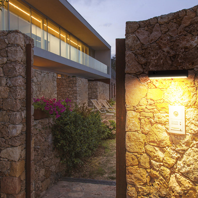 A warm, inviting evening scene of a modern stone villa with a stone pathway leading through an open gate. A black wall-mounted light fixture on the right illuminates a white sign, while the warm glow highlights rustic stone walls, green shrubs, and a lounge chair in the background.