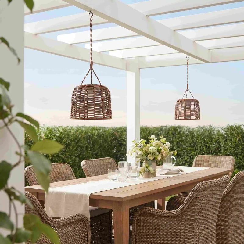 Two woven wicker pendant lights hang over a wooden dining table on a white pergola in an outdoor patio setting with wicker chairs and greenery.