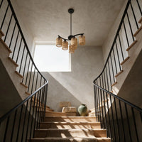 A modern multi-light chandelier with woven rattan shades hangs over a wooden staircase in a textured plaster hallway with black metal railings.