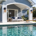 Outdoor patio seating area beside a swimming pool, featuring a white sofa with navy and white striped cushions, a wooden side table, a wooden bench, and a house with blue-gray shingles and white columns in the background.