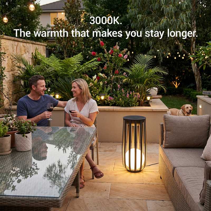 Outdoor patio scene at dusk with a couple sitting at a glass table and a dog resting nearby. A tall cylindrical outdoor floor lamp with a glowing warm light stands on the patio.