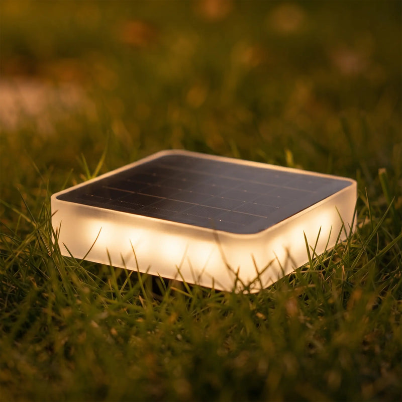 A square solar path light glowing with warm white light on a green grass lawn at night.