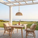 Outdoor dining set on a wooden deck under a white pergola with a view of the ocean. The set includes a rectangular light wood table, two woven rattan chairs with beige cushions, and a hanging woven pendant light.
