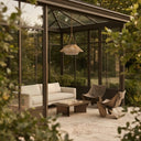 A modern outdoor patio area enclosed by a glass conservatory with dark metal framing, furnished with a white sofa, two sculptural brown chairs, a low wooden coffee table, and a woven pendant light. The scene is framed by soft-focus green foliage in the foreground.