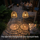 Two illuminated rattan pendant lights casting shadows on a wooden deck with people sitting at a table in the background.