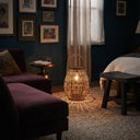 A woven rattan lantern-style floor lamp with a warm glowing light inside, placed on a textured rug in a styled bedroom with dark blue walls, a bed, and a dark purple sofa.