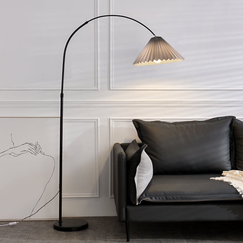A modern black floor lamp with a curved arm and pleated shade, shown lit next to a dark gray sofa in a minimalist room with white paneled walls.