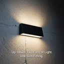 A slim rectangular black solar wall light mounted on a textured gray wall, casting warm light upward and downward. White text at the bottom reads: 'Up.