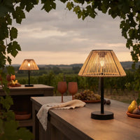 A modern table lamp with a black metal base and a woven natural-fiber shade glowing with warm light, set on a wooden table in an outdoor vineyard at dusk.