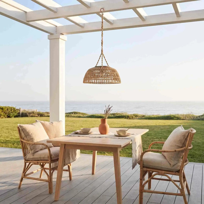 Outdoor dining set on a wooden deck under a white pergola with a view of the ocean. The set includes a rectangular light wood table, two woven rattan chairs with beige cushions, and a hanging woven pendant light.