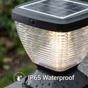 Close-up of Nexi Solar Pillar Light PT0112 ribbed clear dome glowing warm white in rainfall, 5W monocrystalline solar panel on top, water droplets on base, IP65 Waterproof icon overlay