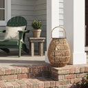 A large, egg-shaped rattan lantern with a rope handle sits on a red brick step, featuring a black solar panel lid and a visible filament bulb inside. In the background, a dark green Adirondack chair with a white cushion is placed next to a rustic wooden side table holding a potted green plant, all set against the light gray siding of a house with white columns.