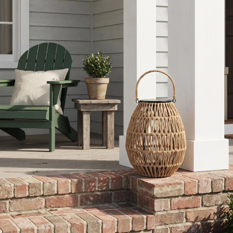 A large, egg-shaped rattan lantern with a rope handle sits on a red brick step, featuring a black solar panel lid and a visible filament bulb inside. In the background, a dark green Adirondack chair with a white cushion is placed next to a rustic wooden side table holding a potted green plant, all set against the light gray siding of a house with white columns.