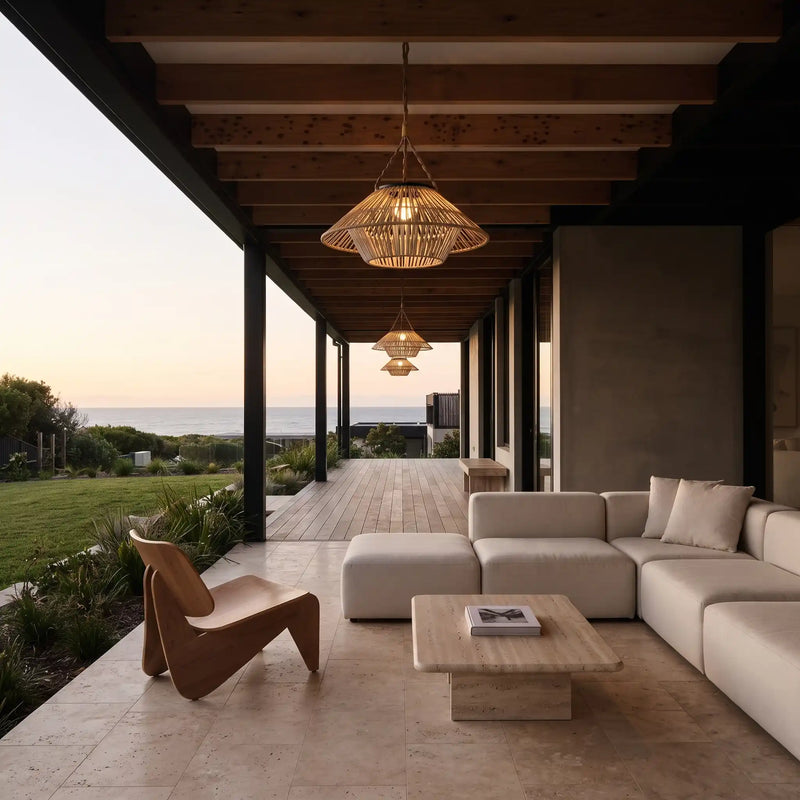 At dusk, two HAIKU PT0902 Rattan Duo Solar Ceiling Lights glow warmly under a dark wooden pergola overlooking the sea. The layered rattan shades create a dramatic interplay of light and shadow, transforming the outdoor lounge into an elegant, artful extension of the home.