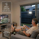 Man relaxing on couch with remote control, solar flood light visible through glass door illuminating the backyard. Three mode cards overlaid: Mode 1 Every Day (dim + motion), Mode 2 BBQ Night (bright all night, 8H timer), Mode 3 Sleep Mode (dimmed for bedroom).