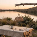A woven wicker pendant lamp hangs from a rustic branch above a table set outdoors by a lake at sunset. The scene includes a linen tablecloth, a metal mug, cutlery, and a folded napkin.