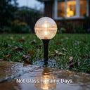 A spherical solar garden light with a glowing warm bulb stands in a puddle on a grassy lawn during rain. The light has a textured clear globe and a black stake.