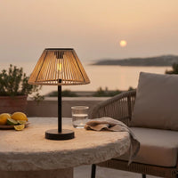 A table lamp with a warm-toned woven shade and black base sits on a stone table outdoors at sunset, with a chair, glass of water, lemons, and a waterfront view in the background.