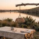 A woven wicker pendant lamp hangs from a rustic branch above a table set outdoors by a lake at sunset. The scene includes a linen tablecloth, a metal mug, cutlery, and a folded napkin.