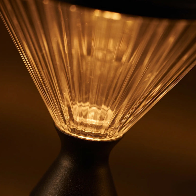 Close-up of a ribbed clear glass pour-over coffee cone with warm amber light glowing through it, mounted on a matte black base against a dark brown background.