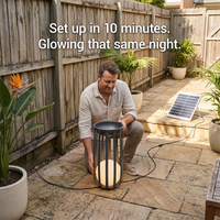 A man kneeling in a backyard patio setting demonstrates a solar-powered outdoor light. The black cylindrical cage-style lamp glows with a warm white light inside, connected to a small solar panel on the patio.