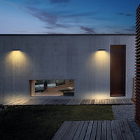 Two modern wall-mounted outdoor light fixtures illuminating a concrete building facade at dusk, with a wooden door, deck, grass, and sky in the background.