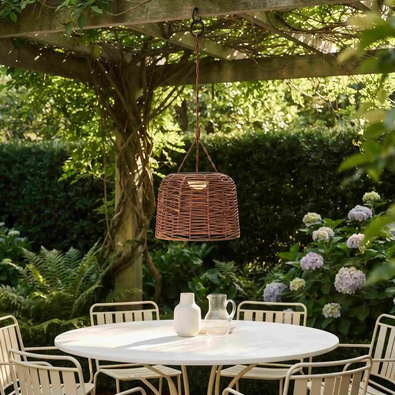 A woven wicker pendant light hangs over a round outdoor dining table in a lush garden patio setting with chairs, greenery, and flowers.