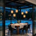 A modern dining room setup under a glass patio enclosure at dusk, featuring a rectangular wooden dining table with a white tablecloth, six black chairs, and five hanging woven pendant lights overlooking a swimming pool.