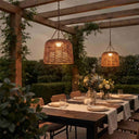 Two woven wicker pendant lights hang over a styled outdoor dining table set with plates, glasses, and floral centerpieces under a wooden pergola at dusk.