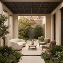 Modern outdoor patio furniture set in a landscaped courtyard, featuring a cream curved lounge chair, two gray upholstered wooden armchairs, a low wooden coffee table, and a wicker pendant light under a dark ceiling.