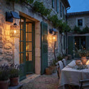 A rustic outdoor wall-mounted light fixture with a woven cage shade and warm glowing bulb, installed beside a stone building with green shutters and a dining table at dusk.