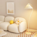 A modern floor lamp with a pleated white shade and curved white stand, glowing warm yellow in a cozy living room setting next to a cream-colored sofa.