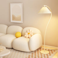 A modern floor lamp with a pleated white shade and curved white stand, glowing warm yellow in a cozy living room setting next to a cream-colored sofa.